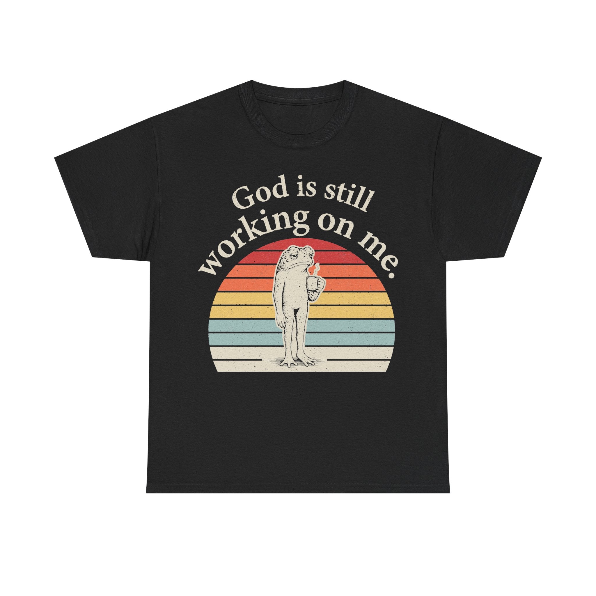 Black T-shirt with retro sunset arc and frog illustration, text God is still working on me