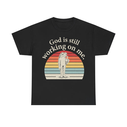 Black T-shirt with retro sunset arc and frog illustration, text God is still working on me